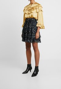 Blusa gialla con volant e tessuto texturizzato, abbinata a una gonna nera a strati con un motivo floreale colorato. Indossata con stivaletti neri.