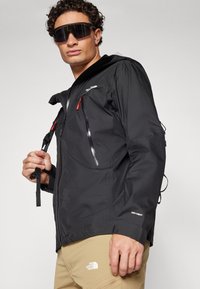 Veste noire imperméable avec zippers rouges et accents réfléchissants. Elle possède une capuche, des poignets ajustables et un logo sur la poitrine et la manche.