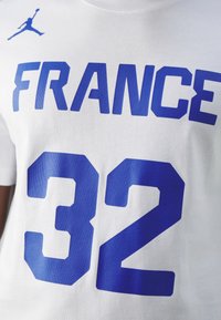 Biała bawełniana koszulka z niebieskimi grafikami: "FRANCE" drukowane pogrubioną czcionką wielkimi literami oraz numer "32" poniżej. Zawiera niebieskie logo Jumpman.