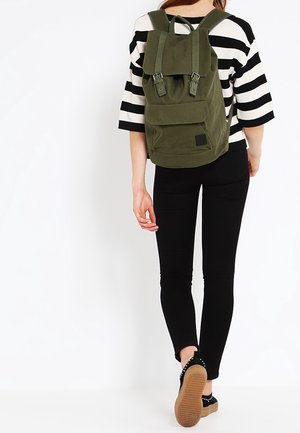 Zelený plátěný batoh s přezkami, přední kapsou a polstrovanými popruhy. Kombinován s černými úzkými kalhotami a balerínami.