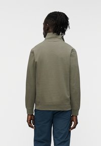 Φούτερ σ ε ελιά πράσινο με ψηλό γιακά, ριμπ μανσέτες και τελείωμα· απαλό ύφασμα· φορεμένο πάνω από μπλε παντελόνι. Απλός, λιτός σχεδιασμός.