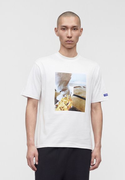 Homme à la tête rasée portant un t-shirt blanc avec une photo imprimée d'une mouette attrapant des frites sur une plage de sable.