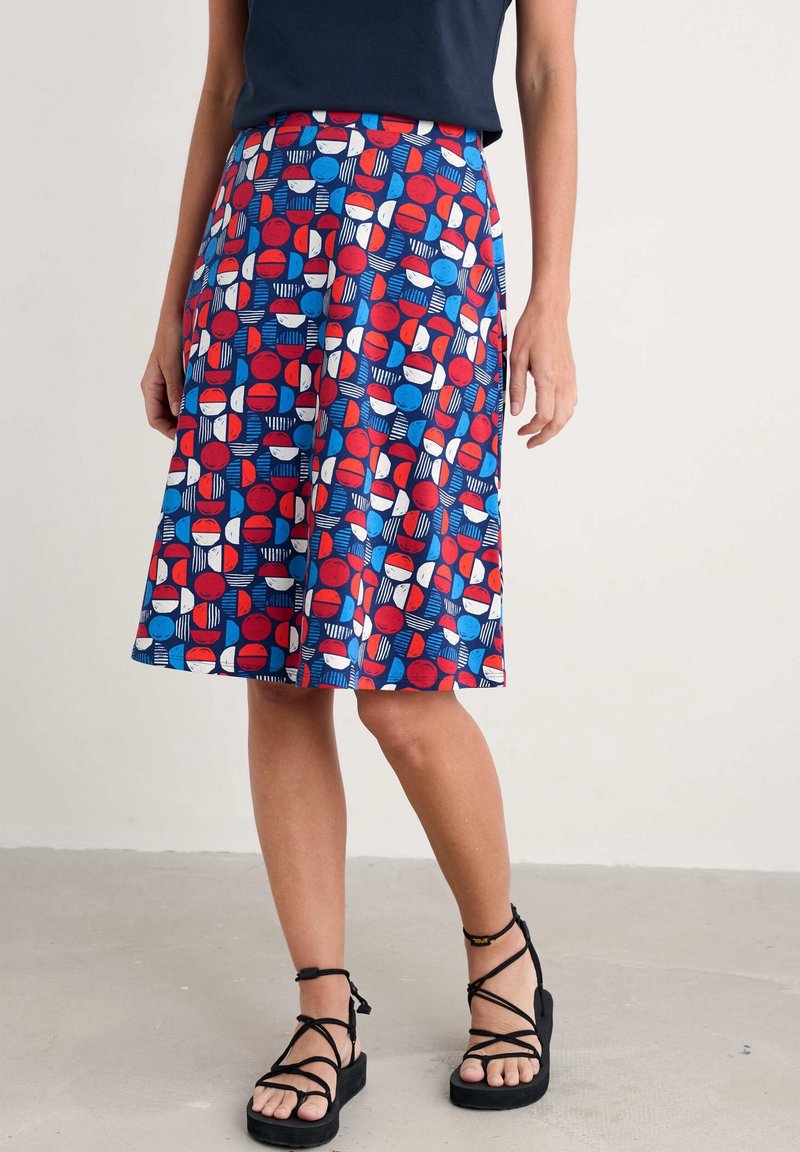 Femme portant une jupe colorée mi-longue avec des motifs géométriques rouges, bleus et blancs, des sandales noires et un haut marine, se tenant à l'intérieur.