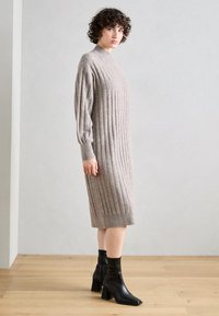 Neule ribbed mekko vaaleassa beigeväri, jossa on korkea kaulus, pitkät hihat ja polven alapuolelle ulottuva pituus. Yhdistetty mustiin nilkkureihin.