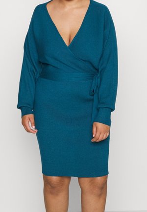 Vrouw draagt een teal geribbelde gebreide wikkeljurk met lange mouwen en een strikceintuur, staand tegen een effen witte achtergrond.