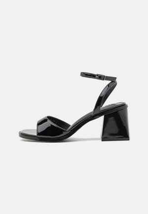 Sandale à talon en cuir verni noir avec un bout carré, un talon bloc, une bride avant ouverte et une bride de cheville avec une petite boucle.