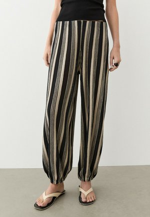 Personne portant un pantalon ample noir et beige à rayures verticales, un haut noir, des sandales beiges, debout devant un fond uni clair.