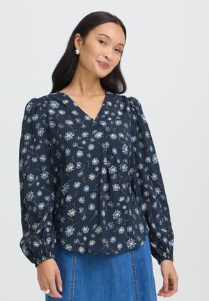 Kvinde med langt mørkt hår bærer en navyblå bluse med hvidt blomstermønster og puffede lange ærmer, kombineret med et blåt denims nederdel.