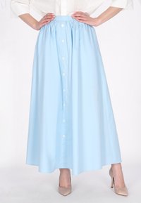 Jupe bleu clair, longueur cheville, fabriquée en tissu lisse, avec une taille froncée et des boutons à l'avant. Porte avec des talons hauts beiges.