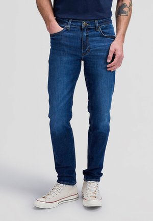 Άνδρας που φοράει εφαρμοστά μπλε τζιν με μπροστινές τσέπες, συνδυασμένα με λευκά πάνινα αθλητικά σε απλό ανοιχτό φόντο.