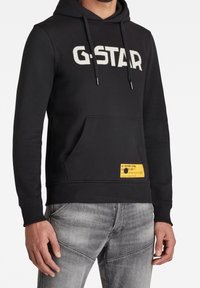 Sudadera negra con un gran logo blanco de "G-STAR", capucha con cordones, bolsillo frontal y una etiqueta amarilla en el lado inferior, combinada con jeans gris claro.