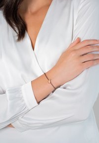 Vékony, barna zsinór karkötő, amelyen egy kis, rózsaarany kutya formájú charm található, egy fehér inggel viselve a csuklón.