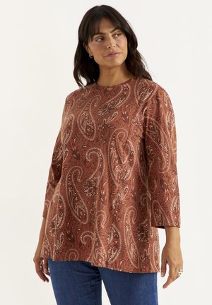 Kvinde med mørkt hår iført en langærmet, brun paisley-mønstret top og blå jeans, der kigger lidt til siden mod en ensfarvet baggrund.
