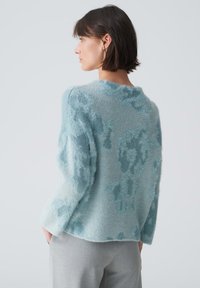 Helleblaue Sweater aus flauschigem Material mit einem strukturierten, abstrakten Muster. Hat einen runden Ausschnitt und eine lockere Passform, getragen über grauen Hosen.
