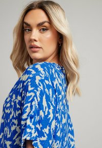 Top stampato blu e bianco con maniche corte, caratterizzato da una vestibilità ampia e un tessuto testurizzato. Include dettagli con bottoni sulle maniche.
