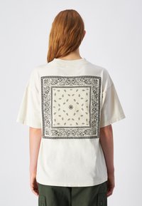 Camiseta de algodón crema con un gran dise�ño de borde paisley negro en la parte posterior, con un patrón central de flores y hojas. Mangas cortas.