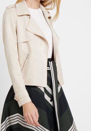 Vrouw draagt een beige suède jack over een wit topje en een zwarte rok met witte en groene diagonale strepen en knopen.