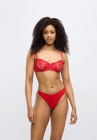 Žena s kovrčavom kosom nosi crveni čipkasti grudnjak i odgovarajuće crvene gaćice visokog reza, stojeći na jednostavnoj svijetloj pozadini.