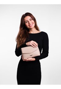 Beige texturovaná kožená kabelka se zavíráním na klopu a embosovaným logem. Modelka drží kabelku a má na sobě černé žebrované šaty.