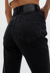 Gros plan sur une personne portant un jean en denim noir taille haute avec des poches arrière visibles et une étiquette de la marque Pull&Bear sur la ceinture.