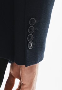 Morska marynarka z mankietem z czterema czarnymi guzikami, fakturowana tkanina i widoczne szczegóły przeszyć. Mankiet lekko wystaje poza nadgarstek.