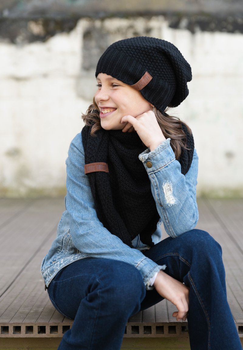 Czarna dziana czapka i szalik, obydwa z teksturowanymi skórzanymi akcentami. Dżinsowa kurtka z przetarciami. Swobodna pozycja siedząca na drewnianej powierzchni.