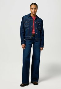 Mørk denimjakke med en strukturerte passform, med to brystlommer og kobberknapper. Kombinert med vide mørke jeans og en rosa topp.