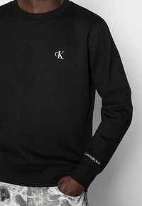 Sweat-shirt noir à manches longues avec logo blanc "cK" sur la poitrine et texte "Calvin Klein Jeans" sur le poignet gauche, porté avec un pantalon gris à motifs.