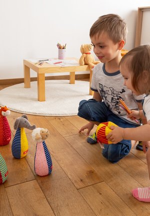 Animali di peluche colorati con motivi e texture diverse sono disposti in una scena giocosa, mentre dei bambini giocano su un pavimento di legno.