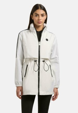 Frau trägt eine weiße Jacke mit Reißverschluss, schwarzen Kordeln und Seitenstreifen, steht vor einem schlichten hellen Hintergrund.