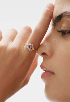 Anillo de oro con una piedra preciosa púrpura, con detalles intrincados alrededor de la piedra; llevado en un dedo cerca de una cara, destacando el diseño del anillo.