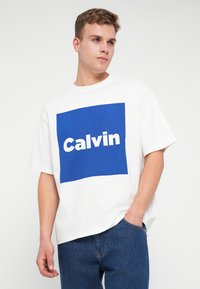 Młody mężczyzna w białej koszulce z dużym niebieskim kwadratem i napisem "Calvin", w połączeniu z niebieskimi dżinsami, pozujący na tle białego tła.