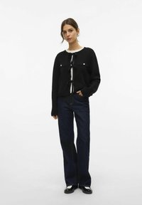 Pull en tricot noir avec un nœud à l’avant, deux poches poitrine et des boutons argentés, associé à un jean bleu foncé à jambes larges et des chaussures noires.