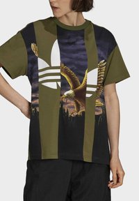 Grön och svart t-shirt med kort ärm som har en stor vit logotyp och en gyllene örn grafik på en mörk bakgrund, med en lös passform.