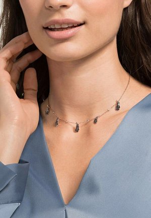 Femme portant un délicat collier chaîne en or avec cinq petits pendentifs noirs en forme de plume, vêtue d'un haut bleu à col en V.