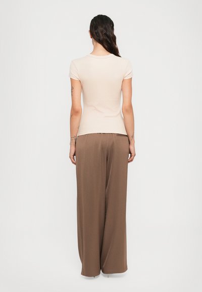 Una donna con longhe capelli scuri che indossa un top attillato beige a maniche corte e pantaloni larghi marrone, in piedi di fronte a uno sfondo bianco semplice.