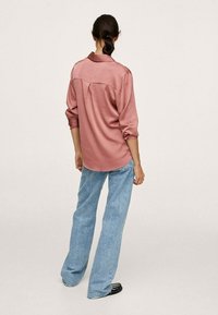 Chemise longue en satin rose à manches longues avec un pli dans le dos, portée avec un jean évasé bleu clair. Des chaussures noires complètent la tenue.