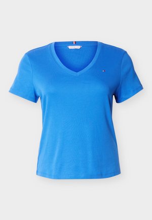 T-shirt bleu à manches courtes avec col en V, logo rouge, blanc et bleu de petite taille sur le côté gauche de la poitrine, présenté sur un fond clair uni.