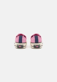 Rosa kanvastennis med blå rand, låg toppdesign, gummisulor och svart etikett med vitt logotyp och rosa stjärndetaljer.