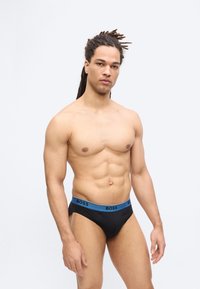 Μυώδης άνδρας με ντραντλόκς που φοράει μαύρα κολάν με μπλε ζώνη μέσης "BOSS", στέκεται μπροστά σε απλό ανοιχτό φόντο.