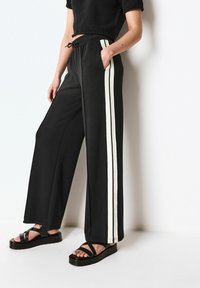 Pantaloni neri a gamba larga con strisce bianche laterali, vita elasticizzata e tasche inclinate; abbinati a sandali platform neri.