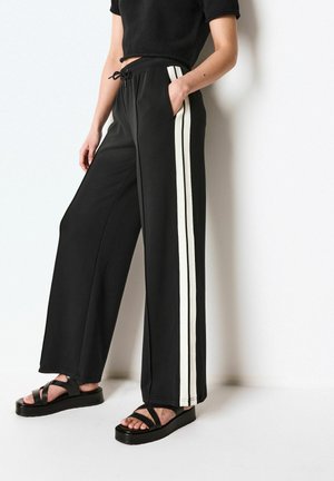 Pantalons larges noirs avec des rayures blanches sur les côtés, taille élastique et poches inclinées ; associés à des sandales à plateforme noires.