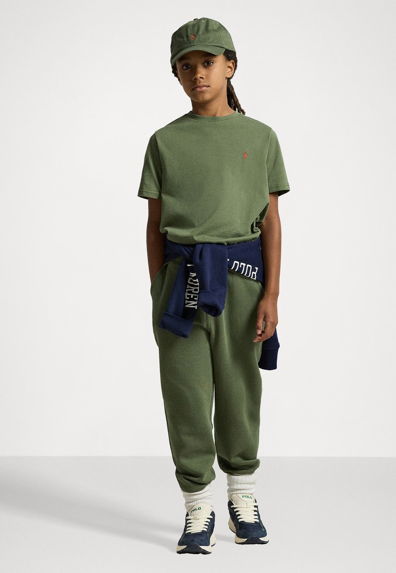 T-shirt verde de algodão com logótipo bordado, calças de jogging a condizer e um sweatshirt azul marinho atado à cintura. Completado com um chapéu verde e sapatilhas.