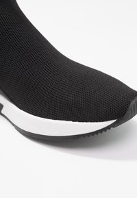 Zwarte gebreide sneaker met een ronde neus en een gestructureerde bovenkant. Voorzien van een witte tussenzool en een zwarte rubberen loopzool met een patroon voor grip.