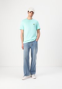 T-shirt en coton vert menthe avec un petit logo de papillon jaune sur la poitrine, accompagné d'un jean large bleu clair et de baskets blanches.