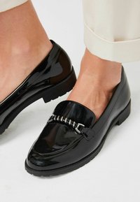 Zwarte lakleren loafers met een lage hak, voorzien van een zilveren kettingaccent over de bovenkant. Glad textuur en een afgeronde neusdesign.