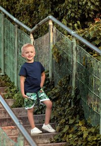 Dziecko ubrane w granatową koszulkę i zielone spodenki w wzory stoi na betonowych schodach obok zielonej metalowej poręczy, otoczone roślinnością.