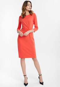 Korallefarbendes knielanges Kleid aus glattem Stoff, mit rundem Ausschnitt, Dreiviertelärmeln und einer verdrehten Frontdetails. Schwarze spitz zulaufende Absätze.