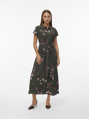 Robe à manches courtes et boutonnée en tissu vert foncé avec un motif floral rose, taille cintrée, jupe ample et longueur mi-mollet. Chaussures à talons brunes.