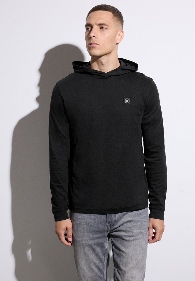 Black long-sleeve hooded shirt with a small logo patch on the chest, paired with light gray jeans, against a plain white background.